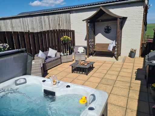 Hot tub on a patio with seating area and a wooden structure against a sunny backdrop.