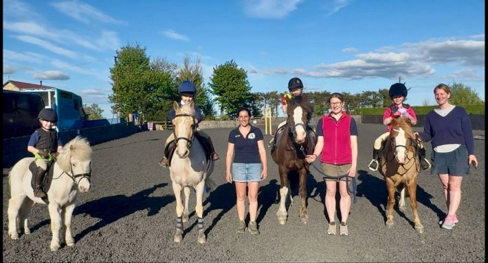 Group of people with ponies standing outdoors on a sunny day.