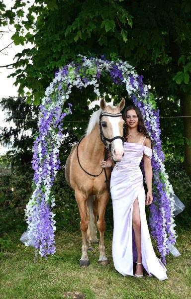 Woman in a lavender gown standing beside a decorated horse under a floral arch.