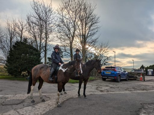 Two riders on horseback near parked vehicles, surrounded by trees and a cloudy sky.